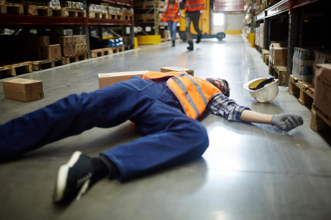 Warehouse Worker Injured on the Job Warehouse worker in an orange safety vest lying on the floor after a workplace accident, representing workers' compensation investigation services in Rancho Santa Margarita, CA.
